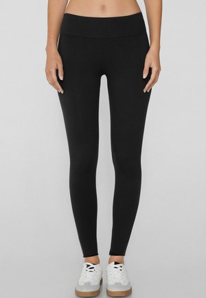 Leggings neri realizzati in materiale elastico con una vita alta. Abbinati a sneaker bianche con accenti grigi. Texture liscia su tutta la superficie.
