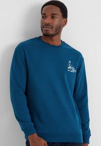 Man wearing a blue long-sleeve sweatshirt with a small white cartoon and text on the left chest, standing against a white wall.