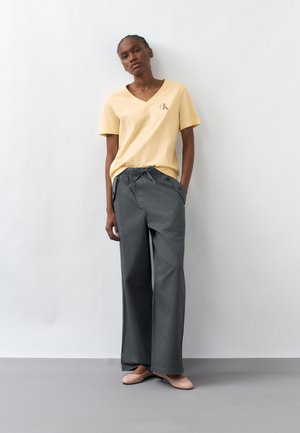 Mujer de pie contra una pared blanca, con camisa amarilla de escote en V, pantalones anchos grises y bailarinas rosa claro, con las manos en los bolsillos.