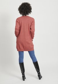 Lång, rostfärgad cardigan i mjukt material, med ribbade muddar och nederkant, kombinerad med blå jeans och knähöga svarta stövlar.