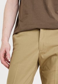 Beige trousers with a smooth texture, featuring a flat front, side pockets, and a fitted silhouette, paired with a brown shirt.