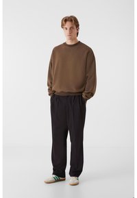 Hanorac maro cu gât rotund, asortat cu pantaloni plisati negri. Modelul poartă adidași crem cu dungi verzi. Ansamblu simplu și casual.