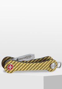 Nyckelhållare i gul kolfiber med texturerad yta, utrustad med en silverfärgad metallklämma och ett rött schweiziskt kors-emblem.