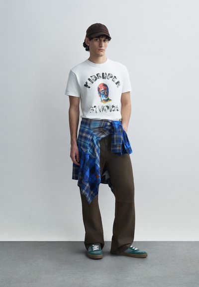 Λευκό T-shirt με πολύχρωμο σχέδιο και κείμενο, μπλε καρό πουκάμισο δεμένο στη μέση, καφέ παντελόνι με φαρδιά πόδια, πράσινα αθλητικά παπούτσια, καφέ καπέλο.