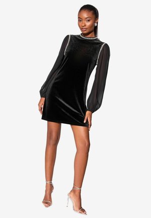 Schwarzes Samtkleid mit durchsichtigen langen Ärmeln, hohem Kragen und verziertem Ausschnitt. Kombiniert mit transparenten Riemchenabsätzen, die die Beine betonen.