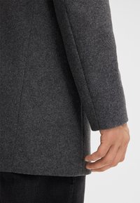 Grau wollener Mantel mit strukturiertem Design. Der Stoff wirkt dick undTexturiert, zeichnet sich durch eine klare, taillierte Silhouette und einen definierten Saum aus.