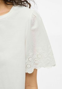 Top blanco de manga corta con un dobladillo de encaje con forma de onda y detalles de cortes, además de suaves mangas abullonadas. Elaborado con una tela ligera.