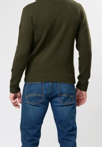 Pull tricot vert olive avec ourlet et poignets côtelés, associé à un jean en denim bleu avec poches arrière standard et coutures visibles.