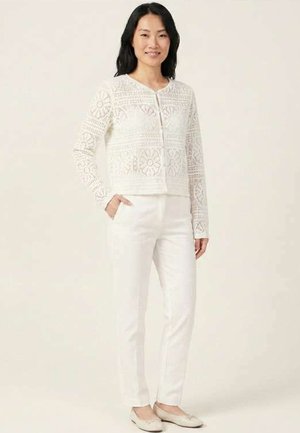 Femme debout portant un cardigan zippé en dentelle blanche, un pantalon blanc slim et des ballerines beiges, une main dans la poche.