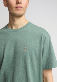 Korte mouwen, T-shirt met ronde hals in gemeleerd groene stof. Voorzien van een klein geborduurd grafisch ontwerp van een tas met een gele print op de borst.