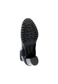 Suela de zapato negra con un patrón de relieve rugoso. El material tiene una apariencia gomosa con surcos y secciones elevadas para mayor tracción. Diseño de tacón bajo.