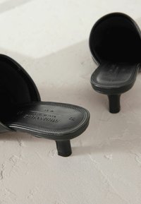 Mules de tacón negro fabricados en cuero suave. Presentan una punta redonda, un tacón bajo y un logo grabado en la plantilla.