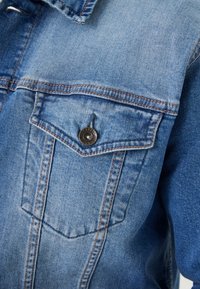 Chaqueta de mezclilla azul claro con cuello puntiagudo, bolsillo en el pecho con botón y costuras visibles. Textura suave.