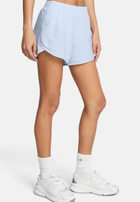 Frau in hellblauen Sportshorts, weißen Socken und weißen Laufschuhen, die vor einem schlichten Hintergrund steht.