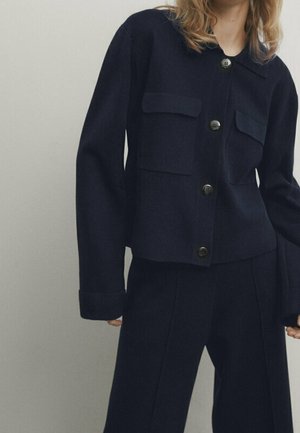 Veste en laine bleu marine avec deux poches poitrine, fermeture à boutons et boutons contrastés. Associée à un pantalon tailleur assorti.