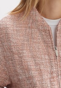 Teksturowana kurtka bomber w delikatnym odcieniu brzoskwiniowym, z zamkiem błyskawicznym, subtelnym wzorem tkanym oraz ściągaczem przy dekolcie.
