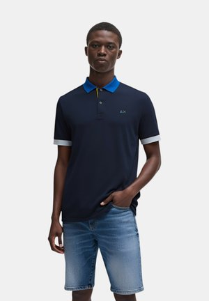 Polo in navy con colletto blu e dettaglio giallo, dotato di un piccolo logo sul lato sinistro del petto, abbinato a pantaloni corti di jeans.
