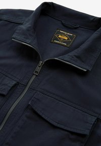 Ciemnogranatowa kurtka z przednim zamkiem błyskawicznym, dwoma kieszeniami z patkami oraz czarno-złotą metką z napisem "Premium Grade 1982 Next Durable Goods" wewnątrz kołnierza.