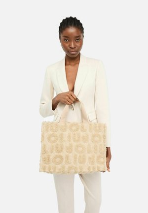 Beige strukturierte Tote-Bag mit hervorgehobenen runden Mustern, aus weichem Material und stabilen Griffen. Getragen von einem Model in einem hellen Blazer.