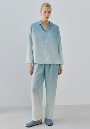 Frau trägt ein locker sitzendes, hellblaues Hemd mit Knöpfen und passende, weit geschnittene Hosen, steht vor einem schlichten hellgrauen Hintergrund.