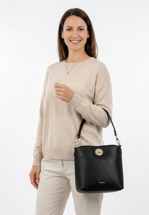 Femme souriante portant un pull et un pantalon beige, tenant un sac à main noir texturé avec un emblème doré et une bandoulière, sur un fond blanc.