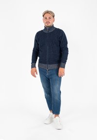 Maglione navy con zip e colletto alto, polsini e orlo a costine in un colore più chiaro a contrasto, abbinato a jeans azzurri e sneakers bianche.