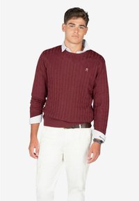 Maglione a maglia cable color bordeaux con scollo rotondo. Abbinato a pantaloni cargo bianchi e una camicia azzurra con colletto, caratterizzata da maniche lunghe e tessuto strutturato.