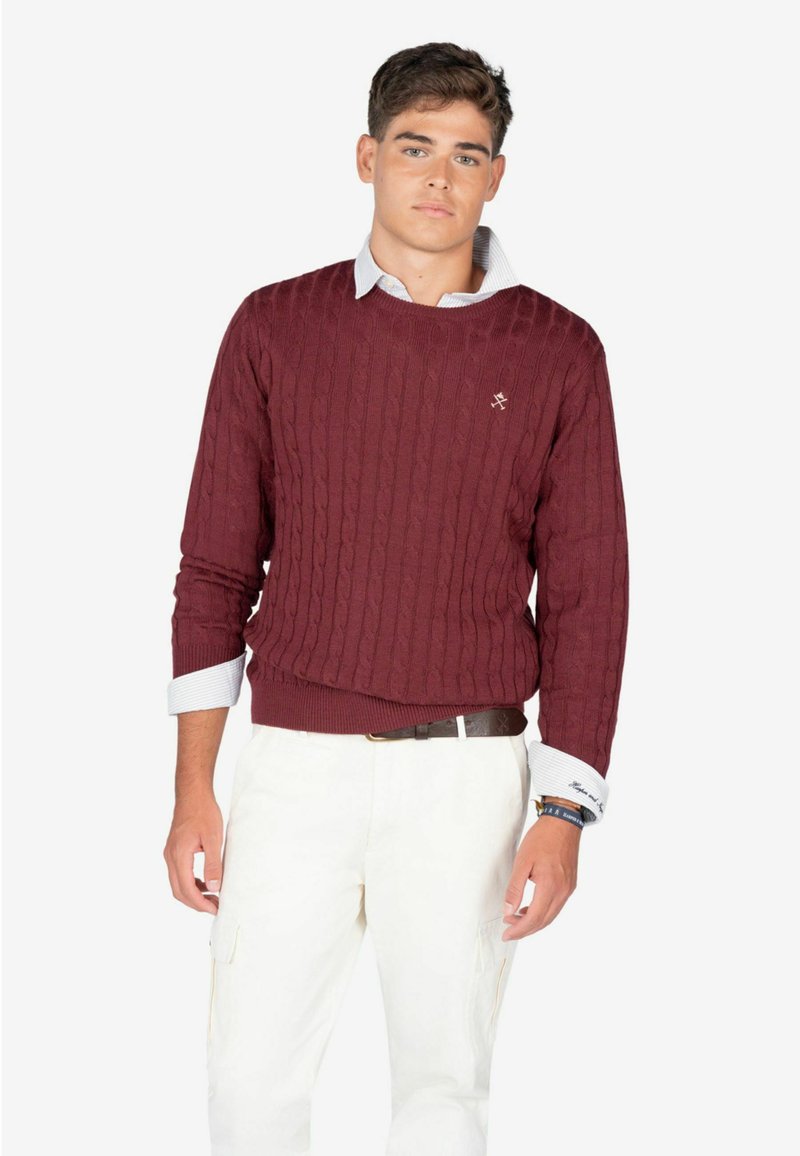 Maglione a maglia cable color bordeaux con scollo rotondo. Abbinato a pantaloni cargo bianchi e una camicia azzurra con colletto, caratterizzata da maniche lunghe e tessuto strutturato.