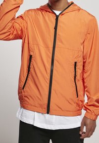 Lättviktsorange jacka med huva, svarta dragkedjor och sidofickor. Tillverkad av ett strukturerat tyg, har den en avslappnad passform och elastiska ärmslut.