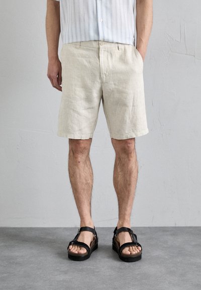 Leinen-Shorts in hellem Beige mit lockerer Passform, ausgestattet mit Seitentaschen und flachem Bund. Kombiniert mit schwarzen Sandalen mit Riemen.