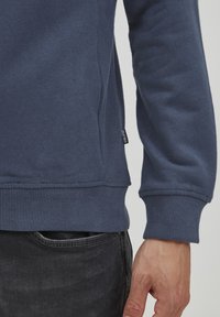 Sudadera azul marino con dobladillo y puños acanalados, costura lateral y una pequeña etiqueta de la marca en un lado inferior. La tela parece suave.