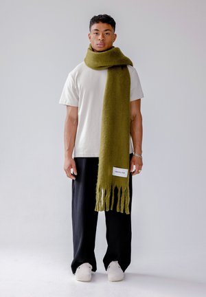 Écharpe vert olive en laine douce avec des bords à franges, étiquetée « ARCTIC FOX », portée sur un t-shirt blanc avec un pantalon plissé noir.