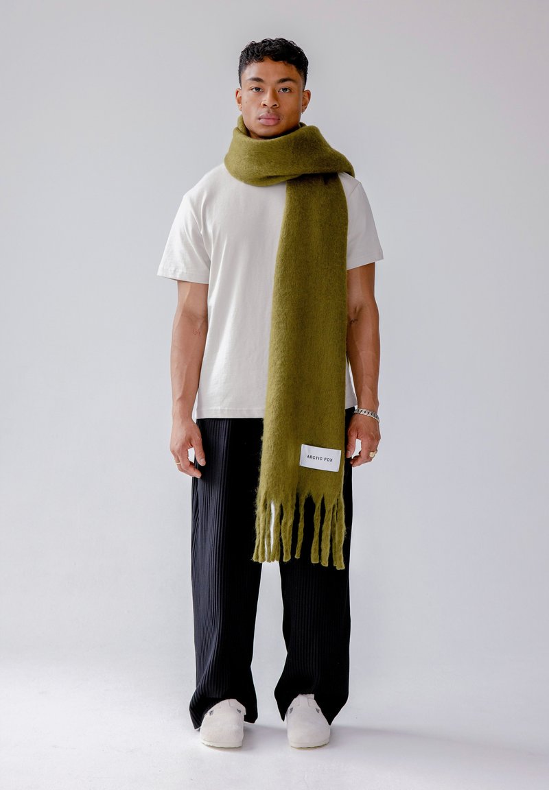 Écharpe vert olive en laine douce avec des bords à franges, étiquetée « ARCTIC FOX », portée sur un t-shirt blanc avec un pantalon plissé noir.