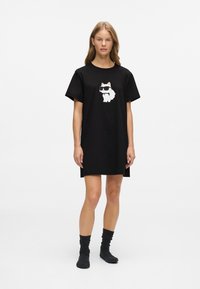 Γυναίκα με μαύρο oversized φόρεμα τύπου T-shirt με γραφικό λευκού γατιού που φορά γυαλιά ηλίου, συνδυασμένο με μαύρες κάλτσες, που στέκεται μπροστά από λευκό φόντο.