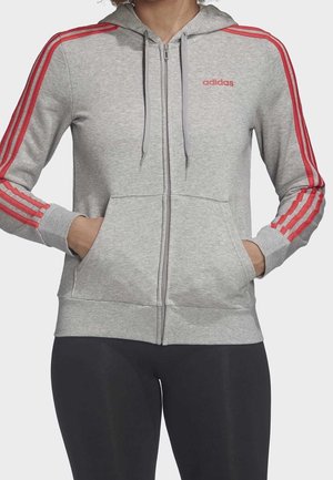 Kvinde iført lysegrå Adidas zip-up hættetrøje med røde striber på ærmerne og rødt logo, med hænderne i forlommerne, kombineret med sorte leggings.