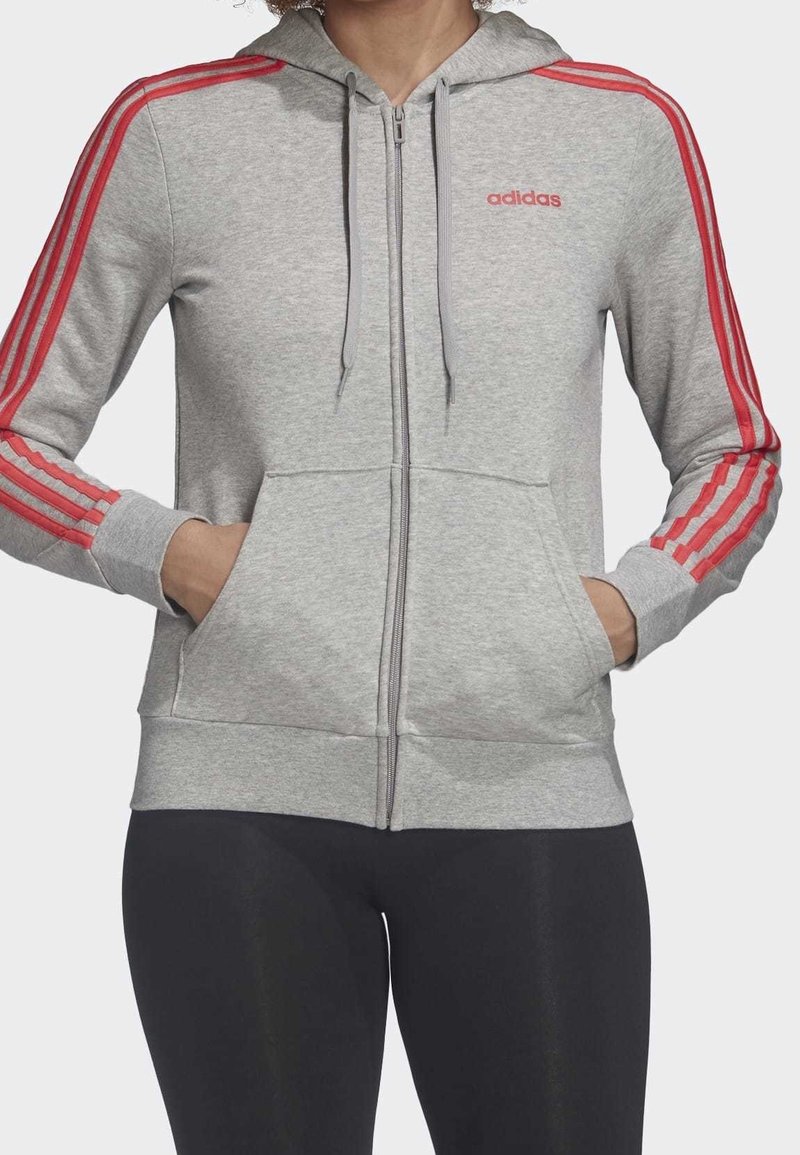 Femme portant un sweat à capuche zippé Adidas gris clair avec des bandes rouges sur les manches et un logo rouge, les mains dans les poches avant, associée à des leggings noirs.