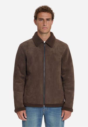 Mann trägt eine braune Jacke mit Reißverschluss, weichem Kragen und Bündchen, kombiniert mit blauen Jeans, steht vor einem schlichten Hintergrund.