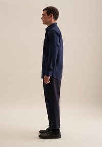 Camicia a maniche lunghe blu navy con colletto abbottonato, abbinata a pantaloni scuri e scarpe nere. Tessuto liscio con una vestibilità su misura.