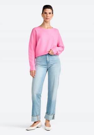 Una donna è in piedi indossando un maglione rosa e jeans chiari, con scarpe bianche. Ha un'espressione neutra e i capelli acconciati, su uno sfondo semplice.
