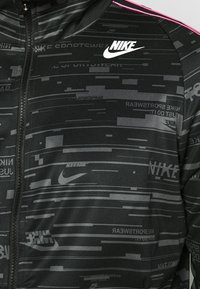 Svart jacka med dragkedja, prydd med en texturerad mönster i olika nyanser och Nike-loggor i grått och vitt, accentuerad med rosa ränder.