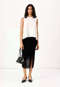 Débardeur blanc sans manches avec ourlet festonné, associé à une jupe midi noire transparente et texturée avec une fente sur le côté. Sac à main noir et escarpins à bout pointu.