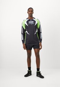Homme portant un t-shirt de sport Venum à manches longues noir et vert, un short noir, des chaussettes noires et des baskets Venum noires, se tenant devant un fond blanc.