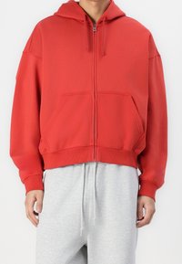 Rote Kapuzenjacke mit Reißverschluss, ausgestattet mit einer Kängurutasche, gerippten Bündchen und Saum. Hergestellt aus weichem Stoff mit lässigem Schnitt, getragen über grauen Sweatpants.