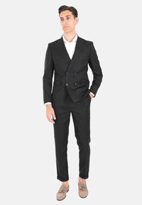 Completo nero doppiopetto con collo a lancia, pantaloni affusolati e camicia bianca, abbinato a mocassini beige.