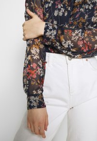 Blommig transparent blus med långa ärmar som har flerfärgade blommor på en marinblå bakgrund, ihop med vita högmidjade jeans.