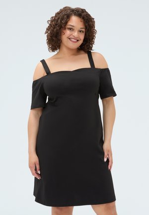 Lachende vrouw met krullend haar, gekleed in een zwarte off-shoulder jurk met dunne bandjes, staand tegen een effen lichte achtergrond.