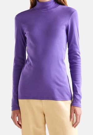Personne portant un haut col roulé violet ajusté à manches longues et un pantalon beige sur un fond clair uni.