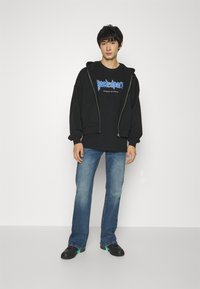 Sudadera negra con cremallera sobre una camiseta gráfica negra, jeans azules y zapatos negros con acentos en teal. Estilo simple, casual y de corte relajado.