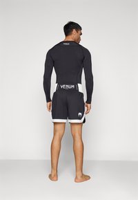 Svart rash guard med lange ermer, kombinert med svarte shorts med hvite detaljer og en logo på midjebåndet. Modellen står barbent.