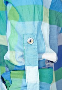 Primo piano di una camicia a quadri blu e verde, con polsino a bottone, tessuto leggero e una texture liscia con una miscela di strisce orizzontali e verticali.